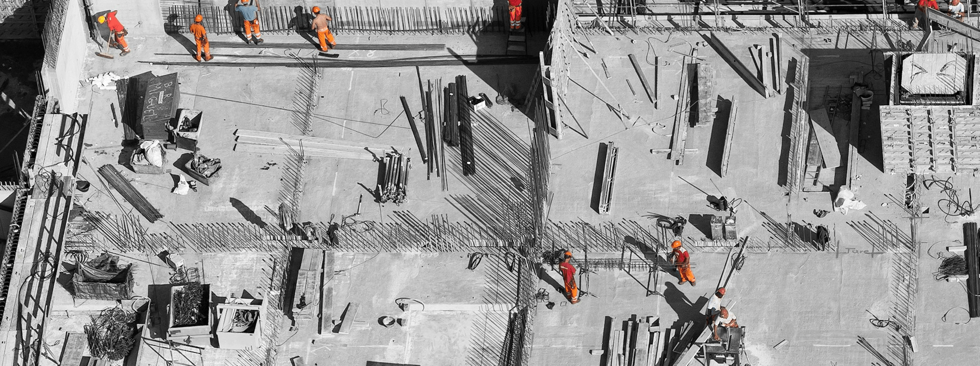 Aerial view of a construction site with workers in high-visibility gear assembling reinforced concrete structures, surrounded by steel rebar, formwork, and building materials.